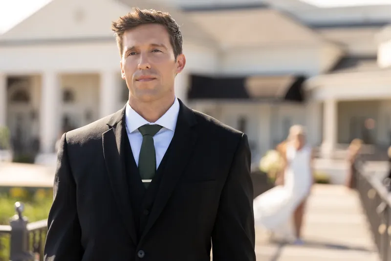 Landon stands in a black suit and blue tie on an outdoor deck at River House Events.