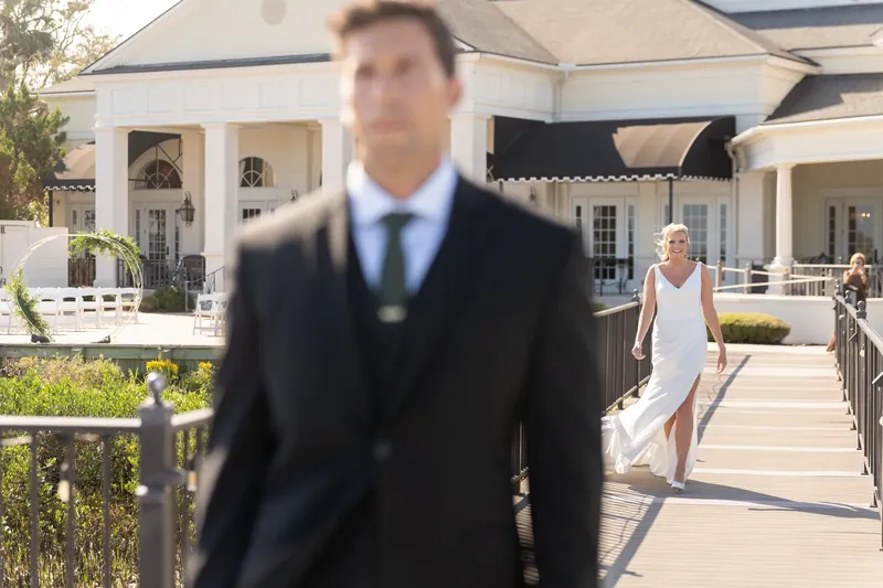 Landon walks across a wooden boardwalk toward Kimberly in a white dress at River House Events.