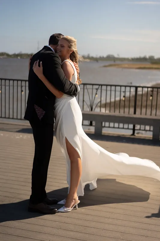 Kimberly and Landon embrace on a waterfront deck with a river and tree line visible behind them at River House Events.