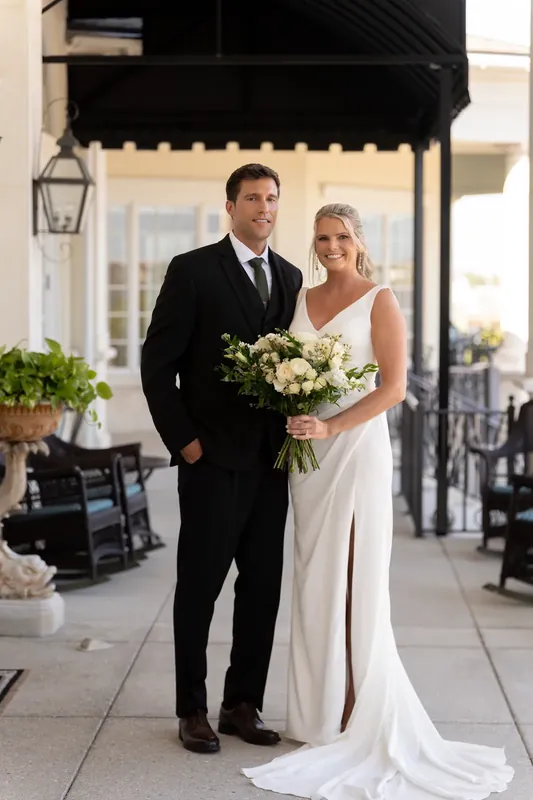 Landon and Kimberly stand together holding a white floral bouquet on a covered patio at River House Events.