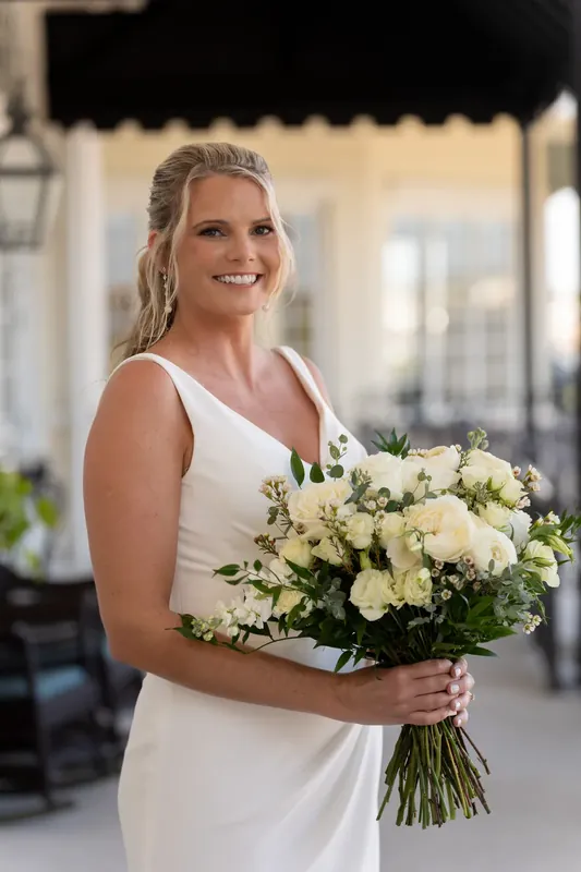 Kimberly holds her white and cream floral bouquet while smiling at River House Events' covered patio.