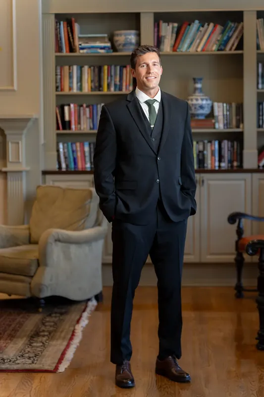 Landon stands in a dark suit with hands in pockets before built-in bookshelves at River House Events.