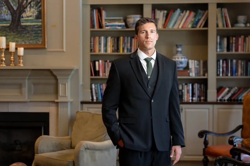 Landon smiles slightly while adjusting his suit jacket in front of a fireplace and library shelving.