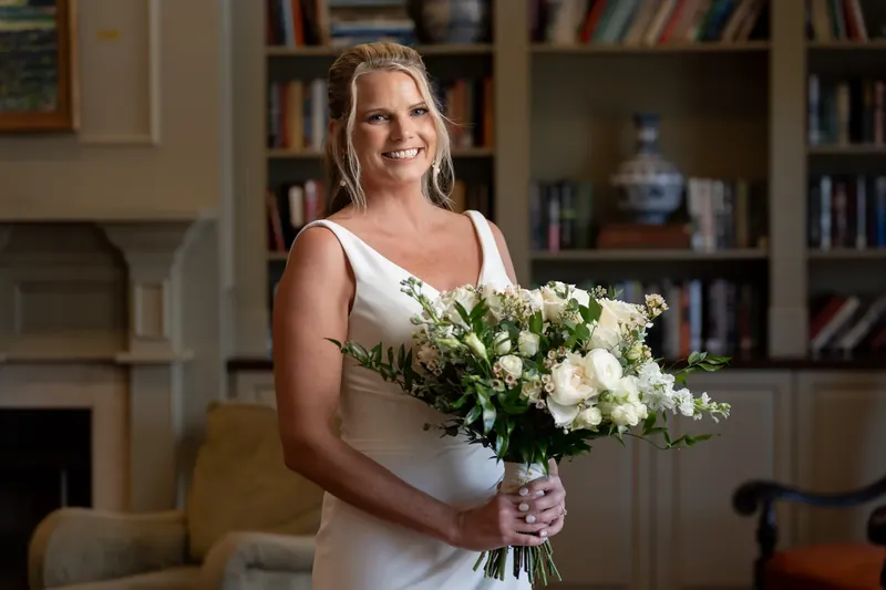 Kimberly holds a white and green bouquet, smiling in her sleeveless wedding dress indoors.