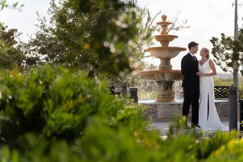 Kimberly and Landon stand near a golden fountain at River House Events, with lush greenery framing the scene.