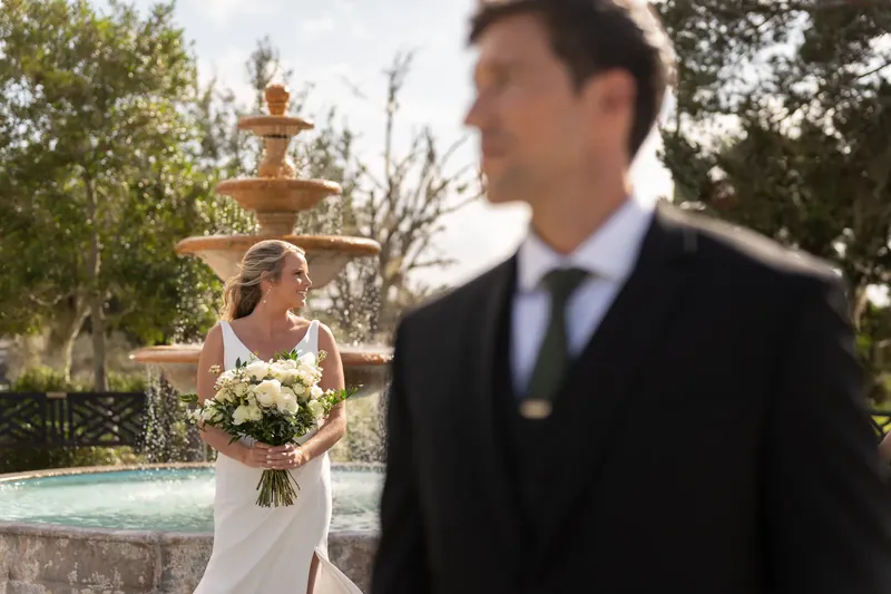 Kimberly holds her white bouquet while Landon faces away, the stone fountain and landscaping in the background.