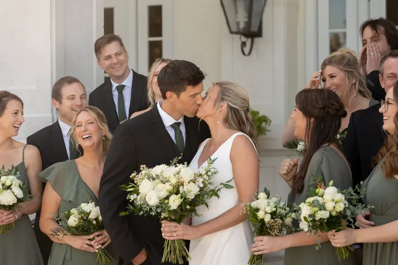 Kimberly and Landon kiss while attendants in green hold white bouquets on the venue's covered entrance.