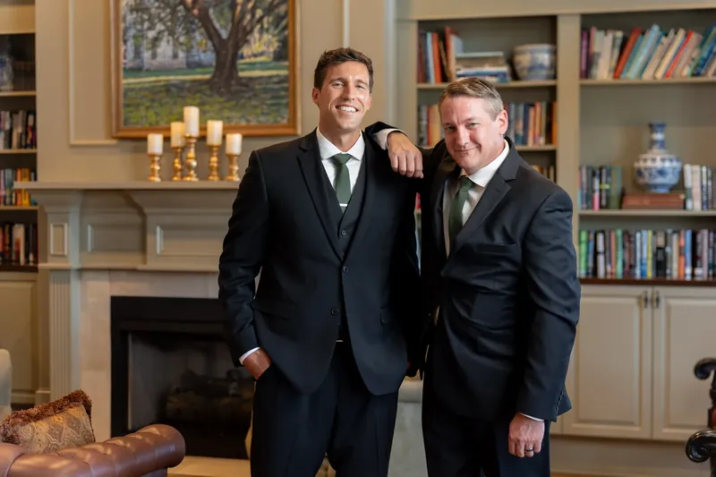 Landon and a groomsman stand shoulder-to-shoulder smiling in the wood-paneled library at River House Events.