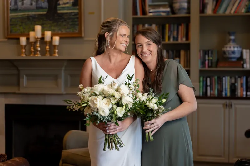 Kimberly holds her white bouquet while posing with a woman in sage green by the fireplace at River House Events.