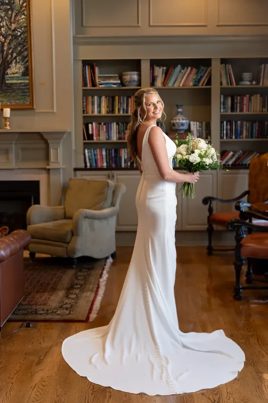 Kimberly stands alone in her sleek white gown with trailing train, holding her bouquet in the library's warm wood interior.