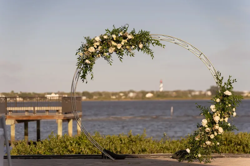 An arched floral arbor draped in greenery and white flowers frames the water and shoreline beyond at River House Events.