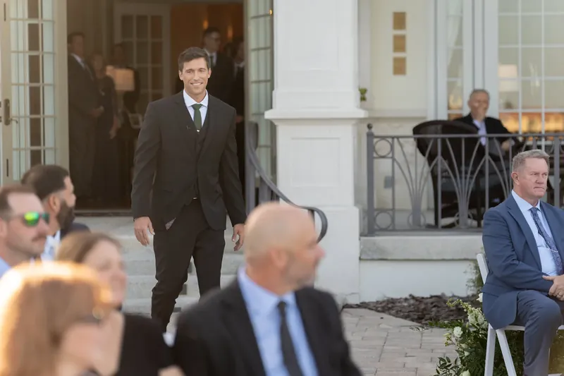 Landon walks through the courtyard toward guests seated on either side of the aisle at River House Events.