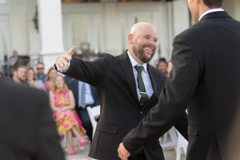 A bearded man in a dark suit and blue tie grins while greeting another guest in formal attire.