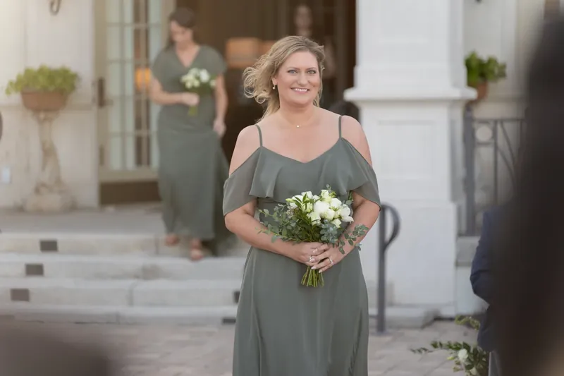 A woman in a gray cold-shoulder dress holds a white bouquet while standing in the courtyard at River House Events.