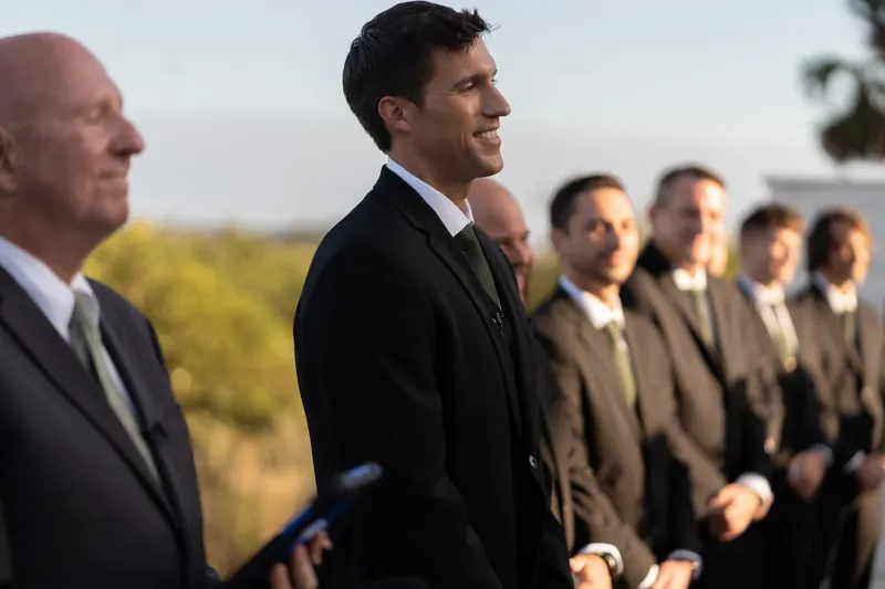 Landon stands with groomsmen in dark suits along a waterfront, smiling while watching the ceremony unfold at River House