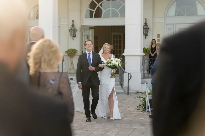 Landon in a dark suit walks beside Kimberly in a white gown down a brick path toward a columned entrance during their first