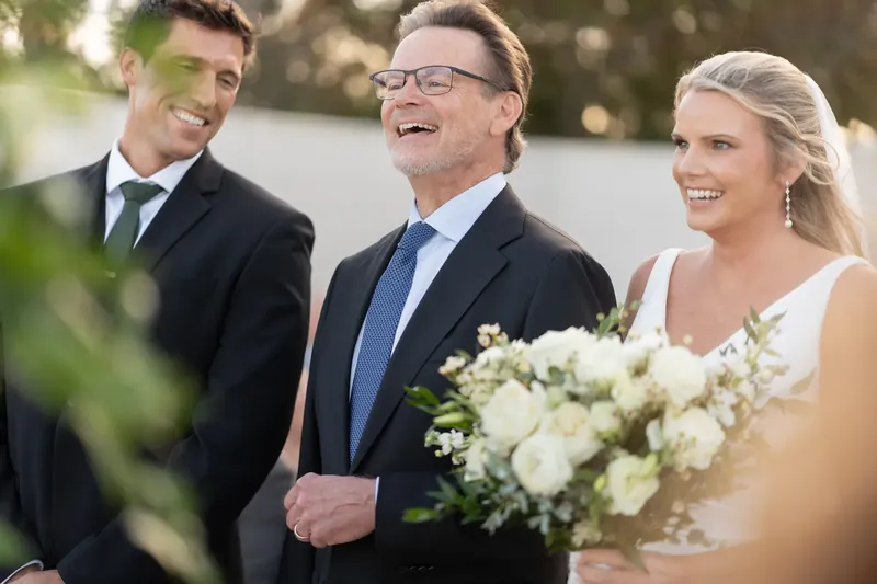 Landon laughs between a man in a blue tie and Kimberly holding white flowers, all three smiling during a family moment.