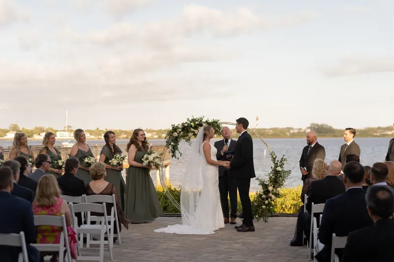 The couple exchanges vows beneath an arch decorated with greenery as guests seated on either side watch their ceremony