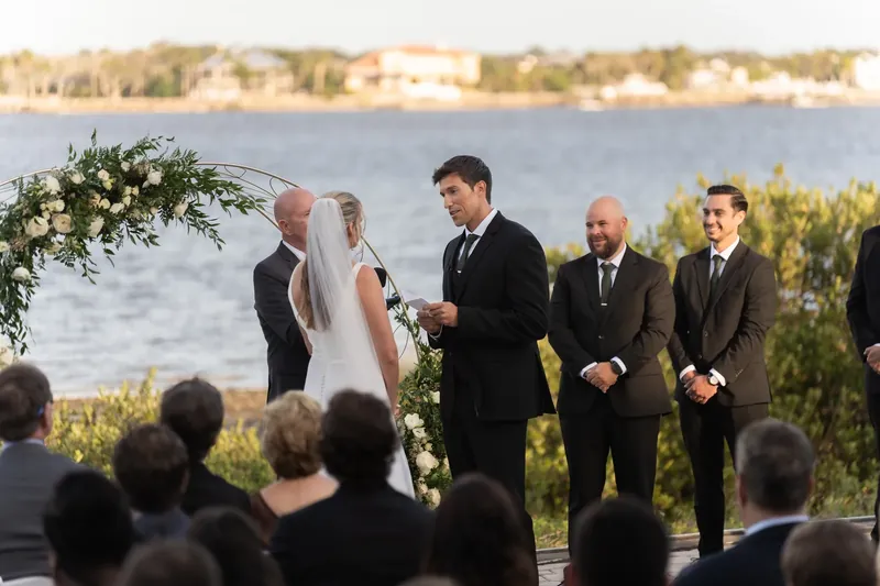 Kimberly in her white gown stands facing Landon at the altar with groomsmen and the river visible in the background behind