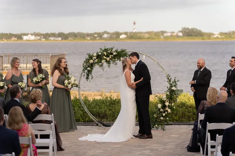 Kimberly and Landon face each other under a circular floral arch while seated guests watch their vows with the water