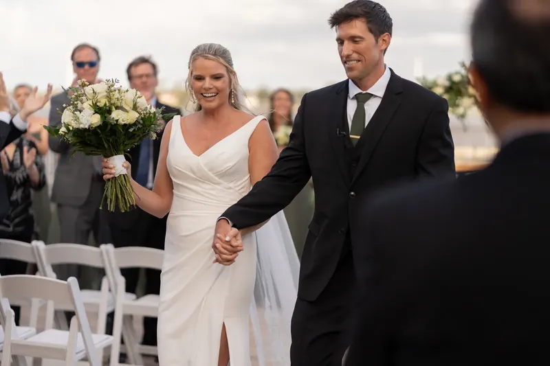 Kimberly and Landon hold hands while walking down the aisle at River House Events, with guests seated behind them