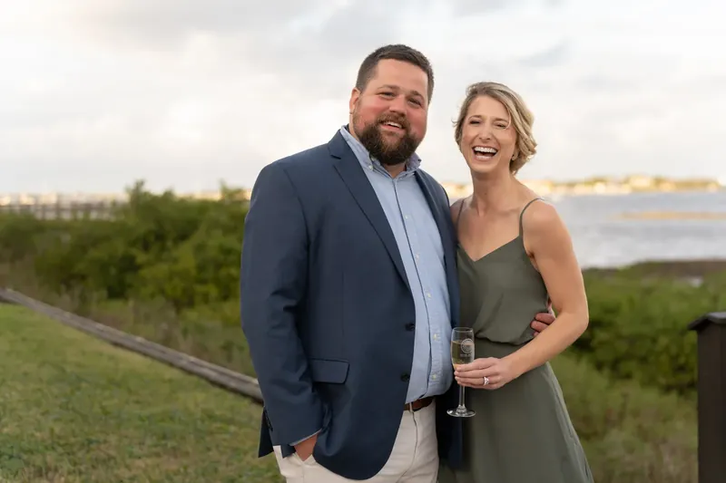 A guest in a sage green dress and a man in a navy blazer smile together with the river visible behind them.