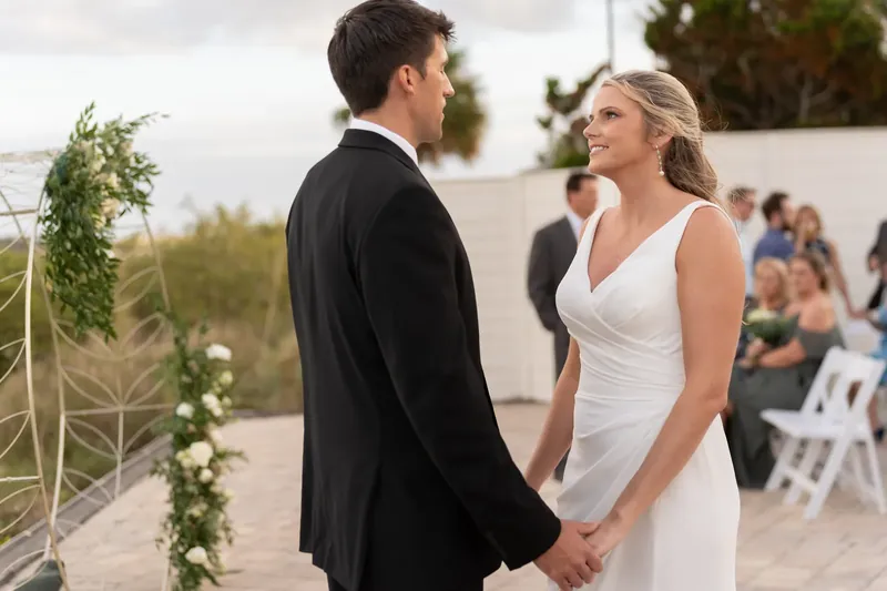 Landon and Kimberly face each other during their ceremony, with guests and a greenery-draped arch visible in the background.