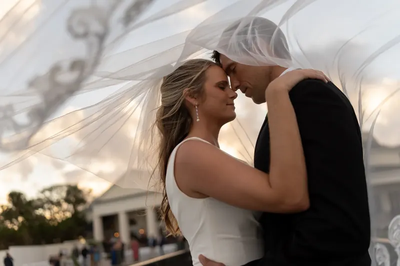 Kimberly and Landon kiss beneath flowing white fabric with the venue's white building visible behind them.