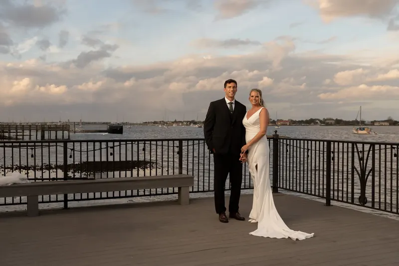Kimberly and Landon stand at a railing overlooking the river, he in a dark suit and she in white.