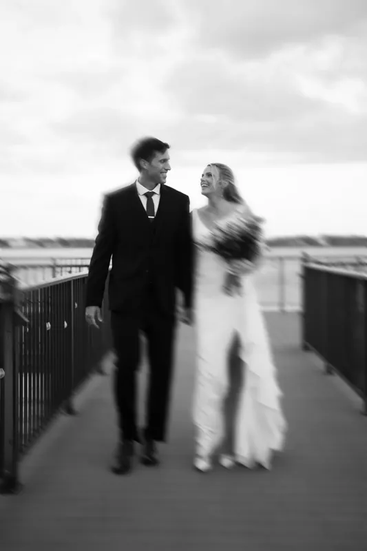 Landon and Kimberly walk together on a waterfront boardwalk in black and white.