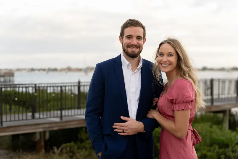 Landon in a navy blazer and Kimberly in a pink dress stand together by a railing overlooking the river at River House Events