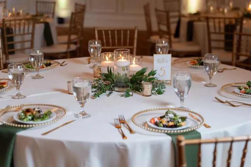 An elegant reception table at River House Events features white linens, place settings with green chargers, and a greenery