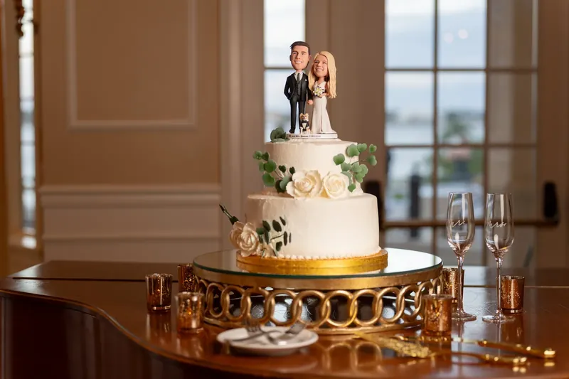 A tiered wedding cake with gold trim and a couple cake topper sits on a gold stand, with windows showing the river view