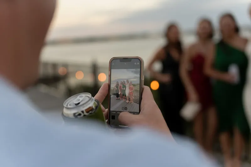 A guest's hand holds a phone to photograph the couple during an outdoor moment on the dock at dusk.