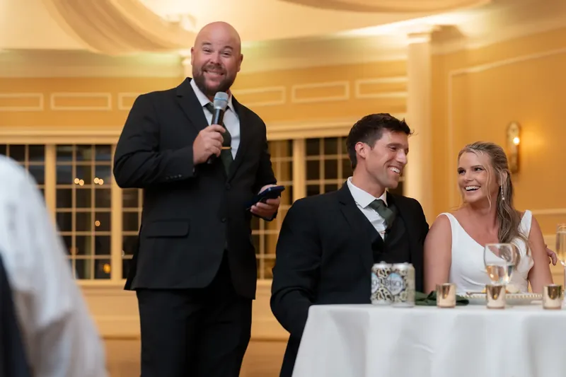 A bearded man speaks at the microphone while Landon and Kimberly listen from the sweetheart table.
