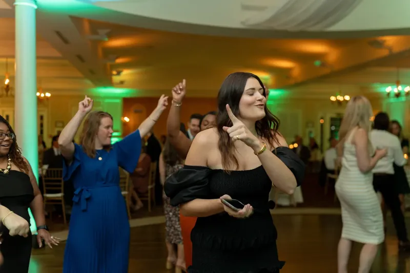 A woman in a black off-shoulder top dances among guests with raised arms during the celebration.