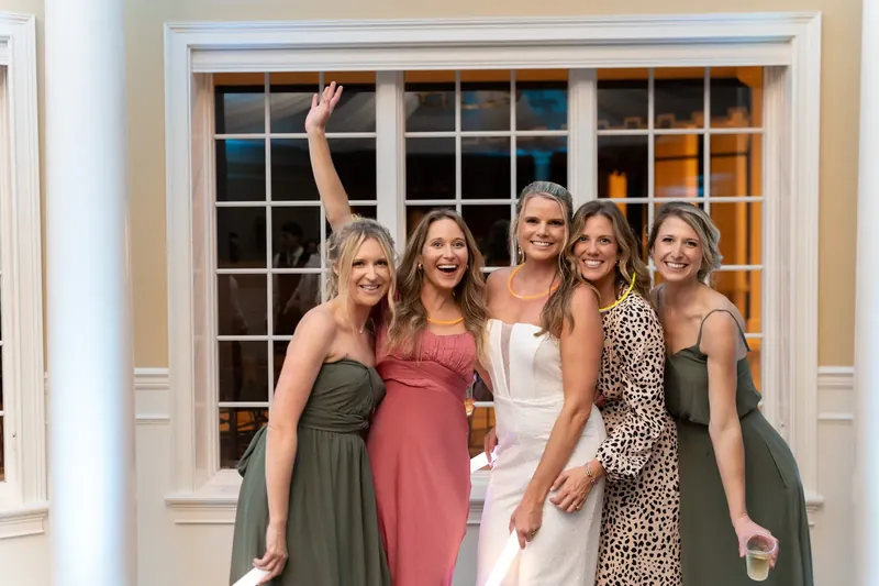Five women in varied bridesmaid dresses wave at a large window during the celebration.