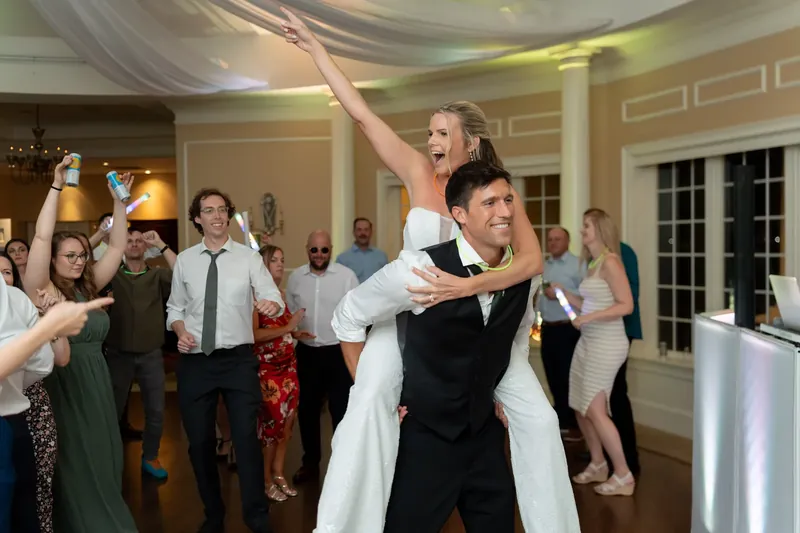 Landon lifts Kimberly onto his shoulders as they celebrate surrounded by dancing guests indoors.