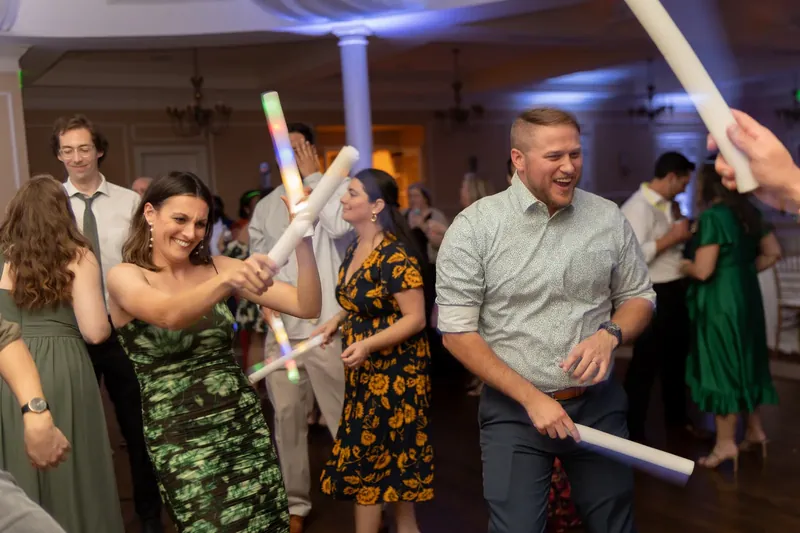 Wedding guests wave white ribbons during a reception game, including a man in a gray shirt and woman in a camouflage dress.