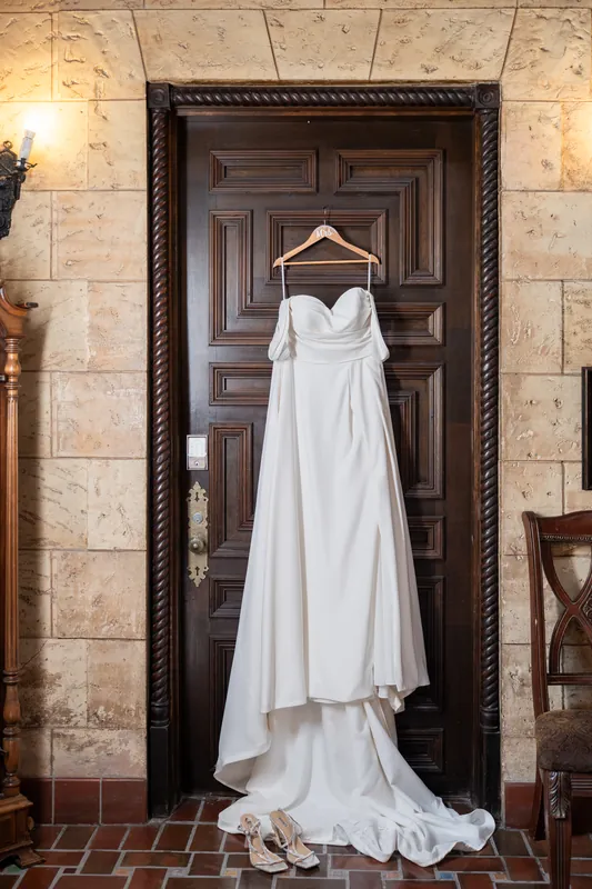 A white wedding gown with a flowing train hangs from a wooden hanger on an ornate dark door.