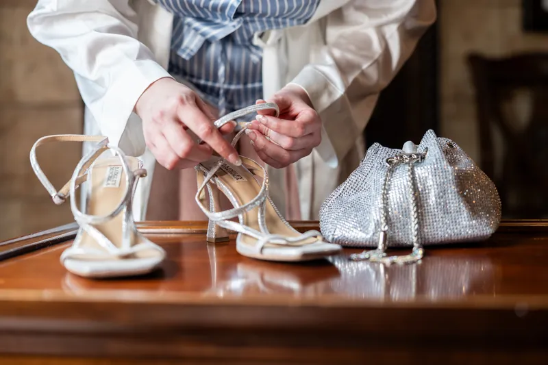 Someone in a striped vest holds white strappy heels and a silver beaded clutch on a wooden table.