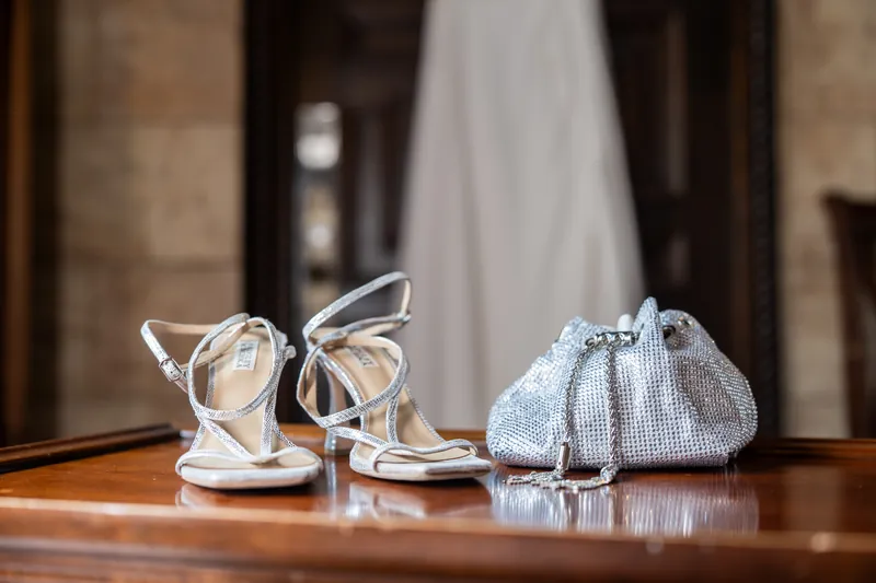 White strappy heels and a silver beaded clutch sit on a polished wooden surface with a white gown visible behind them.
