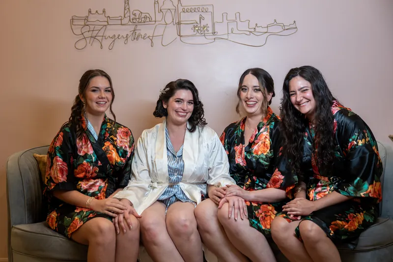 Four people in colorful floral robes sit together on a couch, smiling at the camera in a bridal suite.
