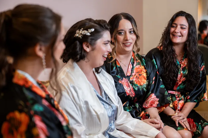 Four people in patterned robes sit together indoors, one wearing a white robe and floral crown in the center.