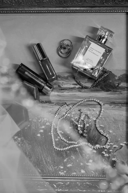 Black and white flat lay of wedding day essentials: perfume bottle, lipstick, mascara, pearl bracelet, and decorative items arranged on ornate fabric.
