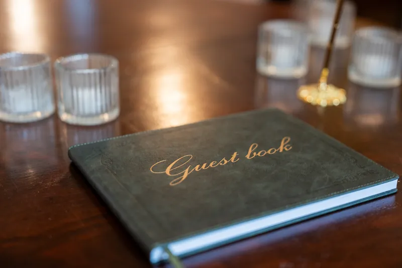 Black leather guest book with gold script lettering rests on a wooden table beside clear glasses at The Treasury on the Plaza.