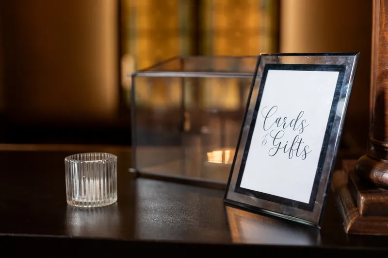 White framed sign reading "Cards & Gifts" stands on a dark table next to a clear glass vase and candlelit background at The Treasury on the Plaza.