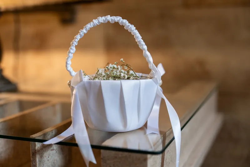 Small white pleated basket with white ribbon handle holds baby's breath flowers on a dark surface at The Treasury on the Plaza.