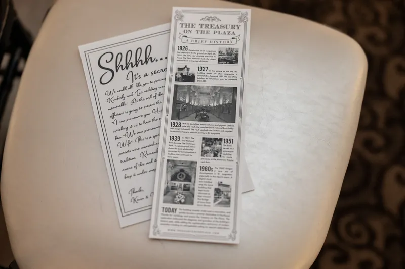 Two printed programs rest on a white round table—one handwritten menu and one vintage newspaper-style layout for The Treasury on the Plaza wedding.