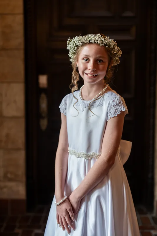 Young girl in a pale blue sleeveless dress with lace detailing and baby's breath flower crown smiles while standing in an arched stone doorway.